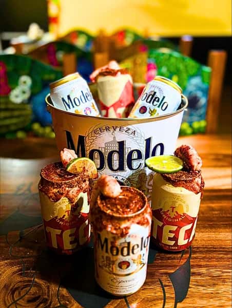 Bucket of Modelo beers with Tecate and Modelo micheladas, garnished with lime, shrimp, and chili seasoning on the rims.