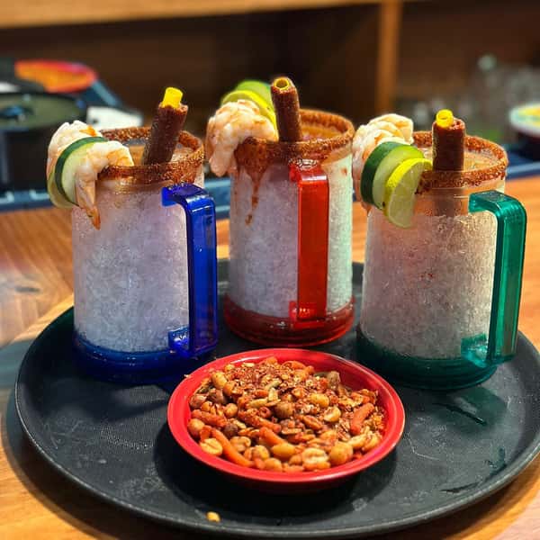 Assorted rimmed beer cans and shots topped with shrimp, lime, cucumber, and fruit, served on a black tray with colorful chairs.