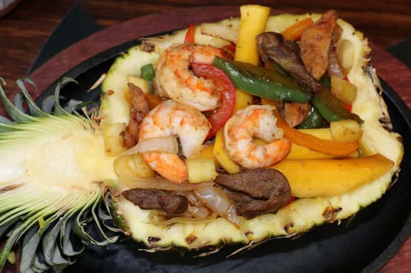 Grilled shrimp, beef, chicken, and bell peppers served in a halved pineapple bowl on a sizzling platter.