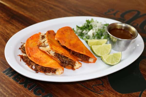 Three birria tacos with melted cheese, served with lime wedges, chopped onions and cilantro, and a side of consommé.