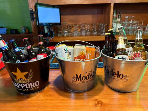 Three buckets of assorted bottled and canned beers including Sapporo, Modelo, Bud Light, Tecate, Corona, and Sol on a bar counter.