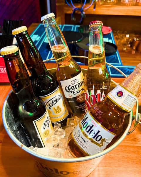 Bucket of ice holding six bottled beers including Modelo, Corona Extra, Sol, and Victoria on a wooden bar counter.