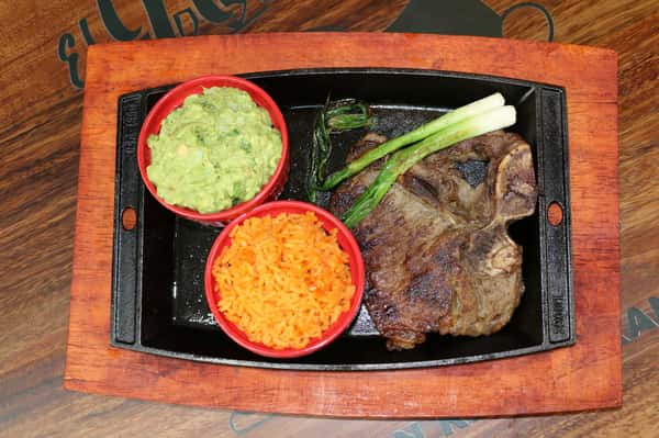 Grilled T-bone steak served with guacamole, Mexican rice, and grilled green onions on a cast iron plate.