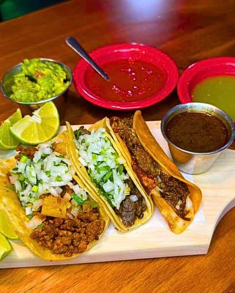 A taco trio platter featuring various meats (likely ground beef, steak, and birria), topped with chopped onions and cilantro. Served with lime wedges, birria dipping sauce, guacamole, red salsa, and green salsa.