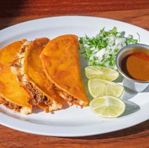 Three birria tacos filled with shredded meat and cheese, served with lime wedges, chopped onions, cilantro, and consommé.