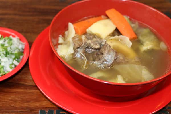 Beef bone soup with carrots, potatoes, and cabbage served with chopped onions, jalapeños, cilantro, and lime wedges.