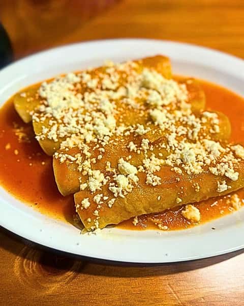 Four enchiladas covered in red sauce and crumbled cheese on a white plate, served on a wooden table.