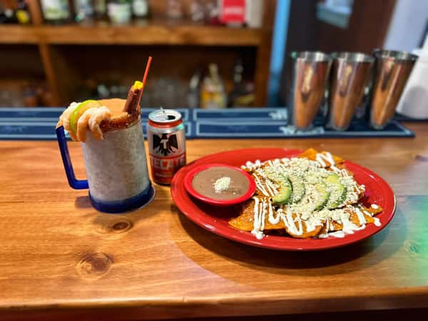 A michelada garnished with shrimp and lime next to a Tecate beer and a plate of chilaquiles with avocado and refried beans.
