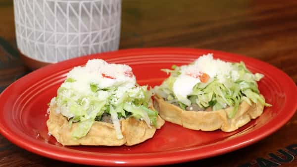 Two Mexican sopes topped with beef, lettuce, sour cream, tomato, and cotija cheese on a red plate.