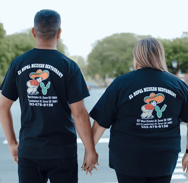 Couple holding hands wearing El Nopal Mexican Restaurant shirts, walking down a tree-lined street.