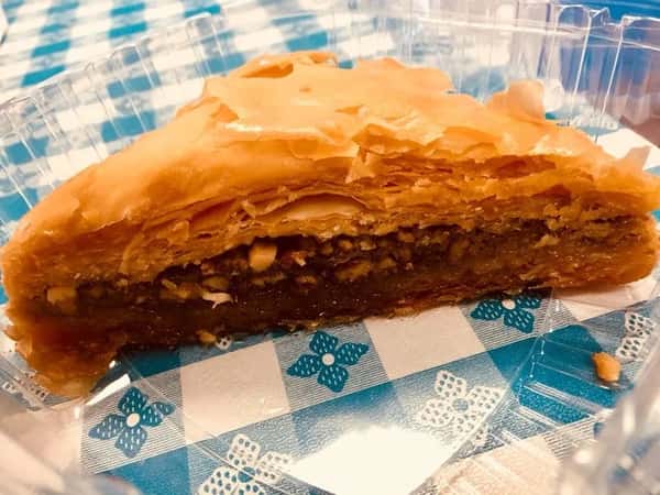 Golden slice of baklava with flaky layers and a sweet nut filling, served in a clear plastic container on a checkered tablecloth.