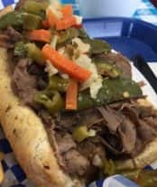 Italian beef sandwich topped with giardiniera on a toasted hoagie roll, served on a blue checkered tray.