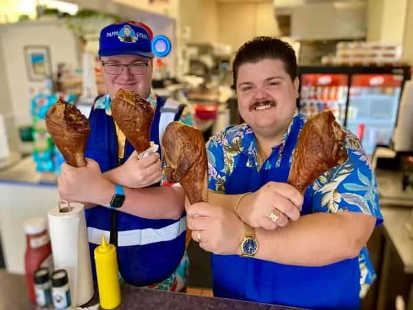 Two staff members at Pappa Gyros smiling while holding large smoked turkey legs behind the restaurant counter.