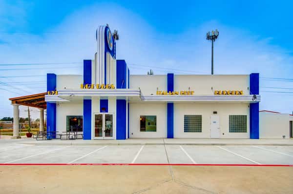This is an image of a Pappa Gyros restaurant building, easily identified by its distinctive blue-and-white color scheme and retro-style architecture. The signage highlights menu staples such as "GYROS," "HOT DOGS," "ITALIAN BEEF," and "BURGERS." The front facade features tall blue pillars, a vertical sign tower with the Pappa Gyros logo, and a clean, inviting appearance with outdoor seating.