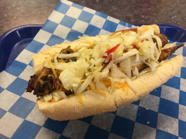 Grilled sausage sandwich topped with shredded cheese and tangy coleslaw, served on a toasted bun with a blue checkered liner.