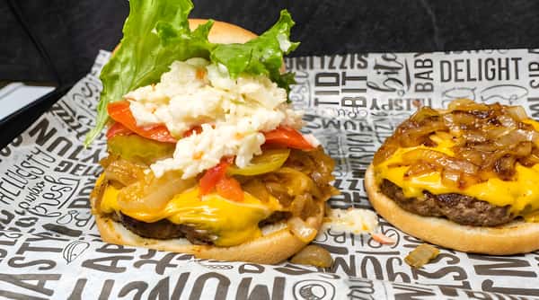 Cheeseburger with melted American cheese, caramelized onions, pickles, tomatoes, coleslaw, and lettuce on a bun.