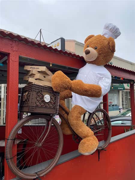Chef stuffed Bear on a bike holding pizza boxes in front of restaurant