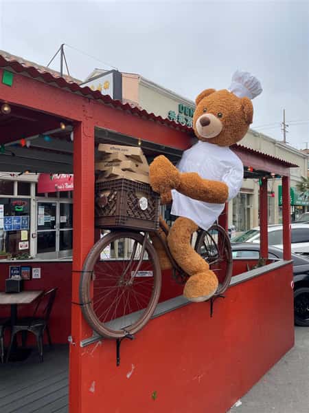 Chef stuffed Bear on a bike holding pizza boxes in front of restaurant