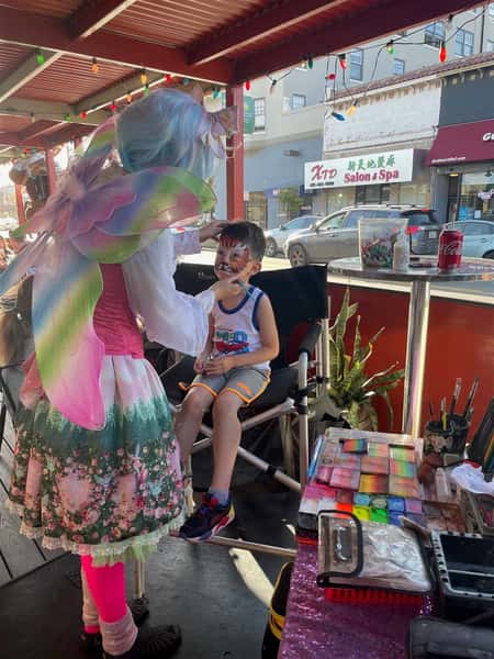 boy sitting down getting his face painted as a tiger from a woman dressed as a fairy