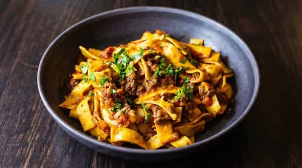 Pappardelle pasta with a rich red sauce, ground meat, and fresh parsley in a dark bowl on a wooden surface.