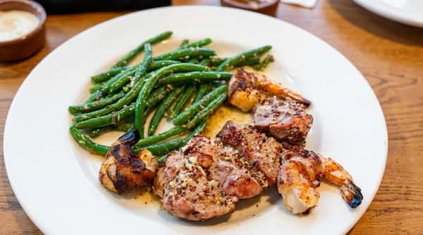 Grilled pork medallions and bacon-wrapped shrimp with sautéed green beans on a white plate.