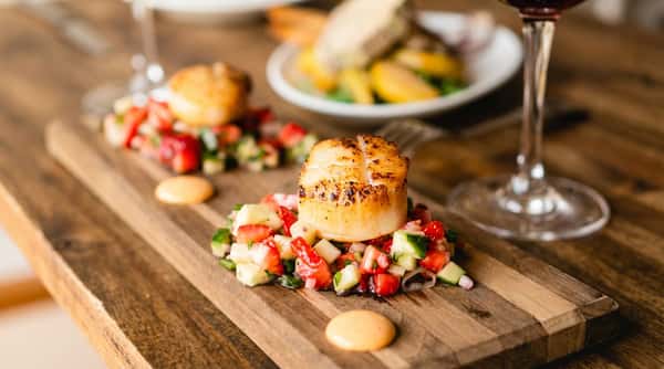 Seared scallops with strawberry and cucumber salsa on a wooden board, with two wine glasses and a blurred appetizer in the background.