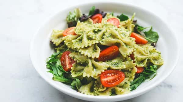 Farfalle pasta coated in pesto sauce with halved cherry tomatoes and green lettuce in a white bowl.