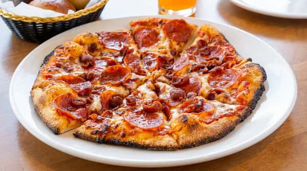 Pepperoni and bacon pizza on a white plate with a drink and bread rolls in the background.