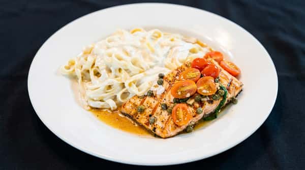 Fettuccine Alfredo with grilled salmon topped with cherry tomatoes and capers on a white plate.