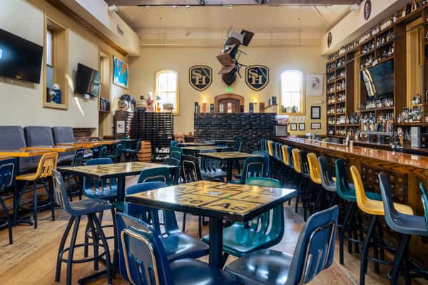 Photo of the "Library Bar" seating area, with high ceilings and high-top tables decorated to look like the Periodic Table of Elements and school chairs converted into bar stools. Photo Credit: The Bacyard.