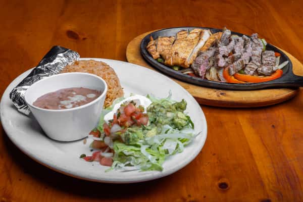 Sizzling steak and grilled chicken fajitas with sautéed peppers and onions, served with Mexican rice, refried beans, guacamole, and pico de gallo for a classic Tex-Mex entrée.