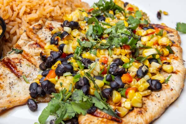 Grilled chicken breast topped with roasted corn and black bean salsa, fresh cilantro, and diced peppers, served with Mexican rice and a side of creamy chipotle sauce, highlighting a vibrant Tex-Mex entrée with bold flavors and colorful presentation.