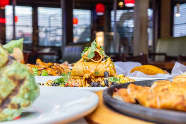 Crispy rolled chicken tacos topped with melted cheese, black beans, corn salsa, and fresh cilantro, served in a lively Mexican restaurant dining room with warm lighting and colorful cantina ambiance.