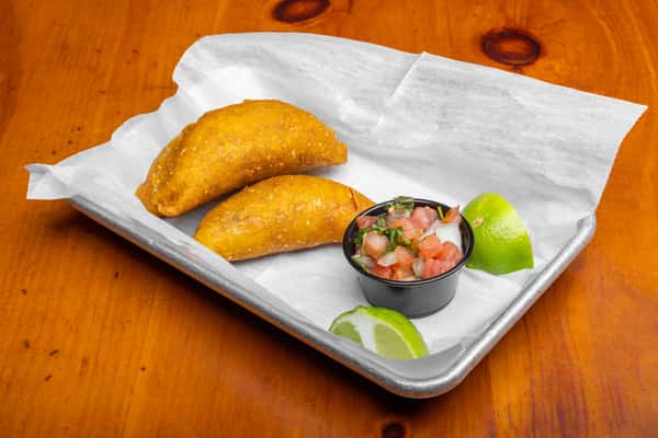 Golden fried empanadas served with fresh pico de gallo and lime wedges on a metal tray, capturing a savory Mexican appetizer with crispy pastry and authentic cantina-style presentation.