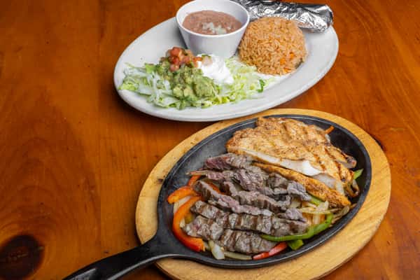 Sizzling chicken and steak fajitas served on a cast-iron skillet with grilled bell peppers and onions, accompanied by Mexican rice, refried beans, guacamole, sour cream, pico de gallo, and a foil-wrapped tortilla, showcasing authentic Tex-Mex cuisine on a rustic wooden table.