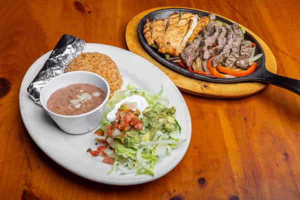 Sizzling chicken and steak fajitas served in a cast-iron skillet with grilled peppers and onions, accompanied by Mexican rice, refried beans, guacamole salad, sour cream, and a foil-wrapped tortilla on a rustic wooden table, highlighting authentic Tex-Mex restaurant flavors.