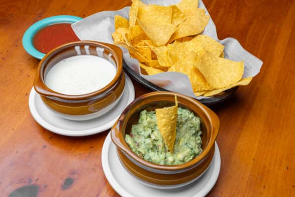 Crispy tortilla chips served with creamy white queso dip, fresh guacamole, and house-made salsa in rustic bowls, showcasing a classic Tex-Mex appetizer spread on a warm wooden table at a lively Mexican cantina.