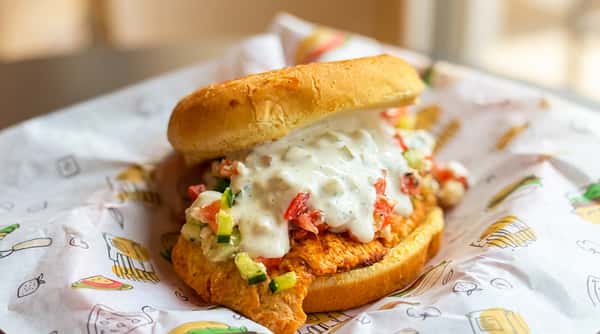 Chicken sandwich on a bun with diced tomatoes, cucumbers, and a creamy white sauce, on patterned paper.