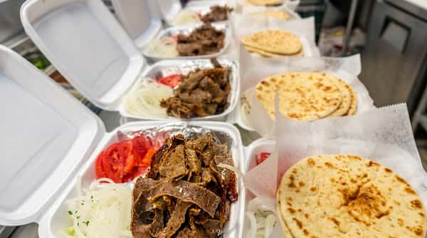 Multiple takeout containers of gyro meat, sliced tomatoes, and onions, with stacks of pita bread.