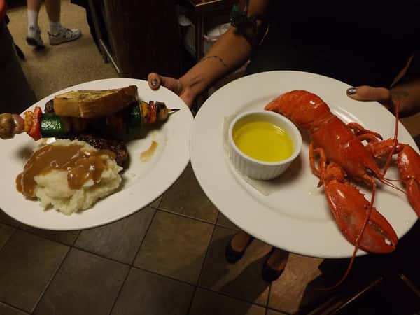 a person holding two white plates. one with mash potaatos and gravy and a shish kebab with a slice of bread. Another plate with a small cup of butter and a lobster