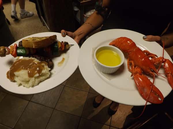 a person holding two white plates. one with mash potaatos and gravy and a shish kebab with a slice of bread. Another plate with a small cup of butter and a lobster