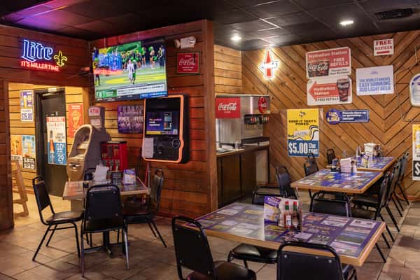 Cozy corner of Darrell's restaurant featuring a drink station, sports-themed posters, neon signage, flat-screen TV with a game, and neatly arranged tables.