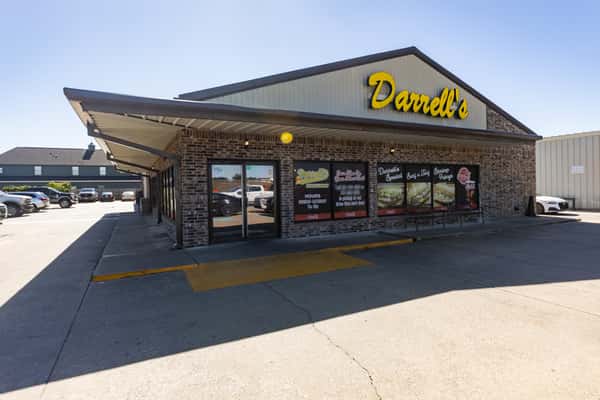 Wide shot of Darrell's po'boy restaurant exterior in bright daylight, featuring a sloped metal roof, large branded windows with food images and hours, and a spacious concrete parking area.
