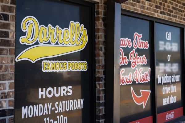 Close-up of Darrell's Famous Poboys window signage on a brick exterior, displaying business hours, drive-thru instructions, and promotional messages in bold fonts.