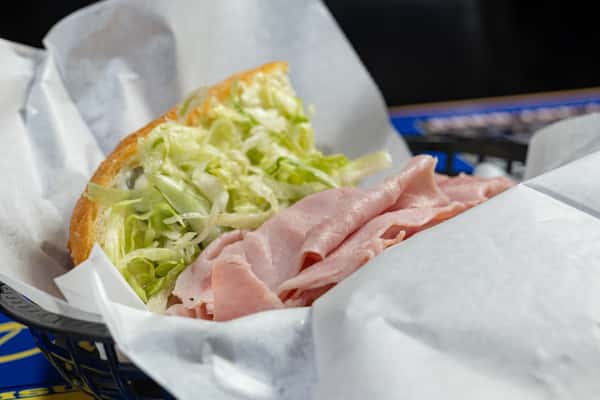 Side view of a ham po'boy sandwich with folded slices of deli-style ham and a generous portion of shredded lettuce on a toasted French roll, served in a black basket at Darrell's.
