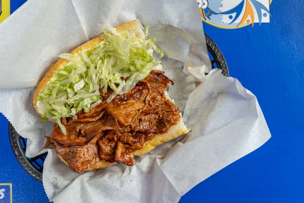 Overhead view of a pulled pork po'boy sandwich with tender barbecue pork, shredded lettuce, and a toasted roll, served in a paper-lined basket on a blue table.