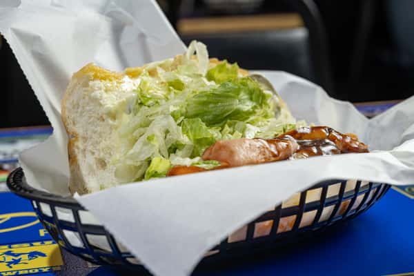 Side angle of a barbecue sausage po'boy sandwich topped with shredded lettuce and glazed sausage slices in a sweet and smoky sauce, served on a fluffy roll in a parchment-lined basket.