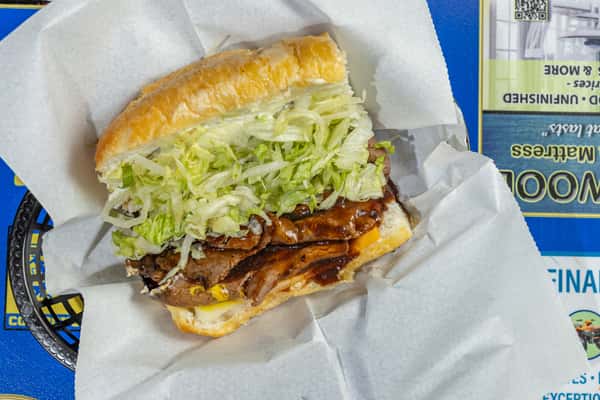 Overhead shot of a Darrell's po'boy sandwich filled with sliced grilled meat, smothered in a rich brown sauce, layered with shredded lettuce and melted cheese, and served in a toasted roll on a parchment-lined basket over a blue branded table.