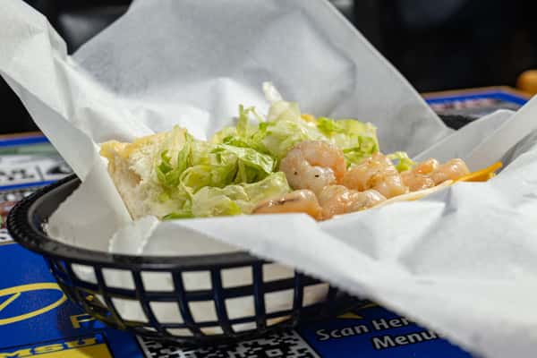Side angle of a Darrell's shrimp po'boy served in a basket, featuring juicy seasoned shrimp and shredded lettuce on a toasted roll, partially wrapped in parchment paper.