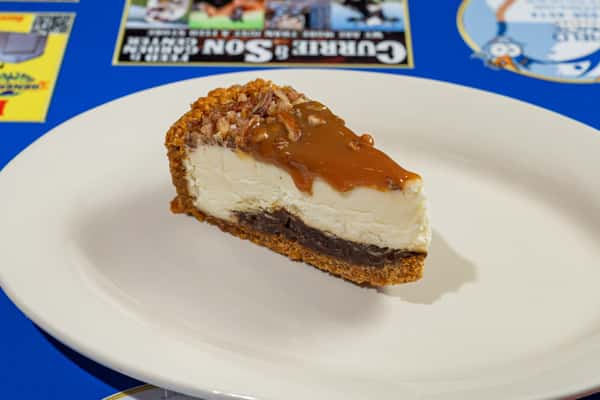 Slice of caramel pecan cheesecake on a white plate, featuring a graham cracker crust, rich chocolate base, creamy filling, and glossy caramel topping with chopped pecans.